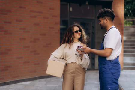 Courier delivering a package to a woman outside a brick building, completing the transaction with mobile payment on her smartphoneの写真素材