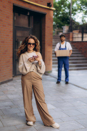 Stylish woman using smartphone while courier delivering package outdoors, modern delivery service and online shopping conceptの写真素材