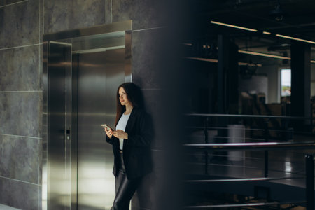 A woman in formal attire standing near an elevator in an modern business office, holding a smartphone, suggesting themes of workplace communication, professional lifestyle, and corporate environments.の写真素材