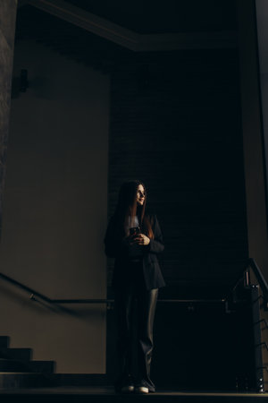 Light illuminates a poised businesswoman in a modern interior space as she holds her smartphone, embodying confidence, technology, and elegance in execution.の写真素材