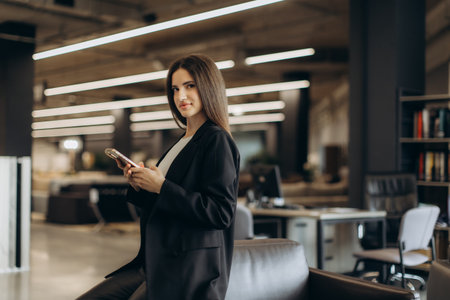 A professional woman wearing a suit holds a smartphone in a contemporary open office environment, conveying productivity and professionalism.の写真素材