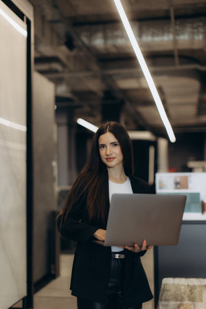 Confident woman in professional attire holding a laptop in a contemporary office. Demonstrates a productive corporate setting and conveys professionalism. Ideal for themes of technology, workplace, and career motivation.の写真素材