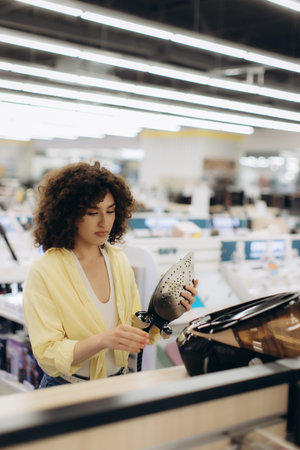 A young woman wearing a yellow shirt carefully examines an iron in a brightly lit electronics store, showcasing product assessment.の写真素材