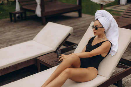 Young woman wearing black swimsuit and sunglasses is relaxing on a beach chair with a towel on her head, enjoying a moment of tranquilityの写真素材