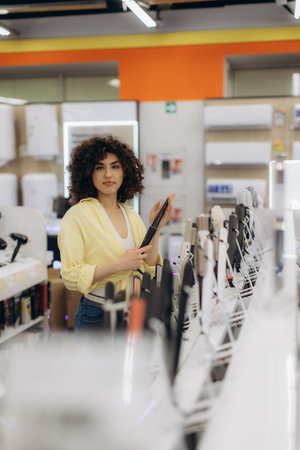 A young woman wearing a yellow shirt examines beauty products on display in an electronics store,...