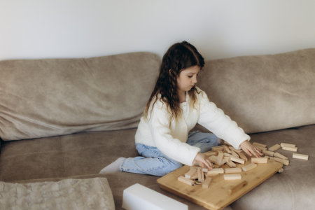 A young girl dressed in casual clothing is sitting on a couch, playing and organizing wooden block pieces, portraying creativity and leisure, in a comfortable home environment.の写真素材