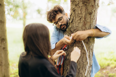 Young couple collaborating, tying a white rope around a tree trunk in a park, showing teamwork and togethernessの写真素材