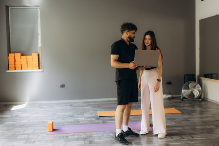 Man and woman in a yoga studio reviewing a fitness program on a laptop. Personal trainer assisting female clientの写真素材
