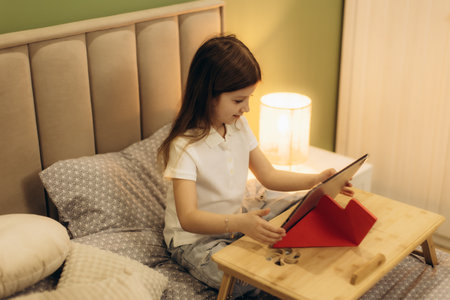 A child comfortably seated on a bed using a tablet for entertainment or learning, illuminated by soft bedside lighting, creating a calm and cozy atmosphere in the bedroom.の写真素材