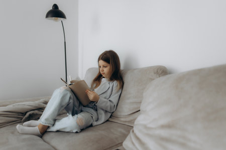 A young girl is sitting casually on a sofa, deeply engrossed in a book. The setting evokes a serene and peaceful ambiance, perfect for moments of relaxation and quiet reading activities.の写真素材