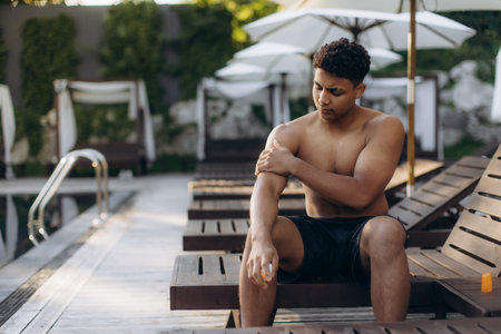 Tourist applying sunscreen lotion on his arm while sitting on a sunbed by the pool, protecting his skin from sunburnの写真素材