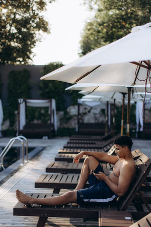 Tourist enjoying summer vacation by the poolside, using his smartphone while relaxing on a sunbed under a parasolの写真素材