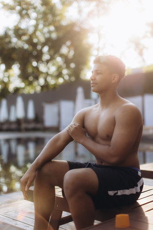 Young man diligently applies sunscreen to his arm, prioritizing sun safety while enjoying a relaxing moment by the poolの写真素材
