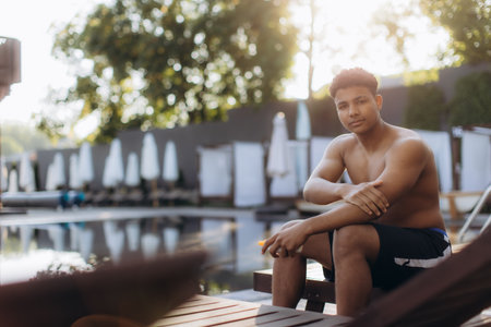 Young man applies sunscreen to his arm while sitting poolside, prioritizing skincare and sun protection during his leisure timeの写真素材