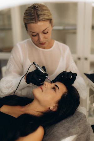 Professional beautician applying permanent makeup to client's eyebrows in a beauty salon for...