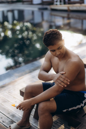 Young man is sitting poolside, diligently applying sunscreen to his arm, ensuring protection from the sun's raysの写真素材