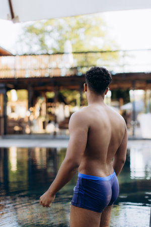 Rear view of a muscular man wearing swim shorts, standing by the poolside of a resort, contemplating a swimの写真素材
