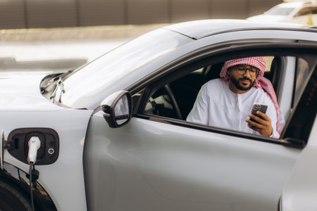 Arab man sitting in an electric car, charging the vehicle and using a smartphone, connecting to green modern technologyの写真素材
