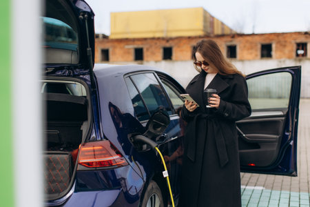 Woman standing next to electric car, charging vehicle at station, texting on phone while holding coffee cupの写真素材
