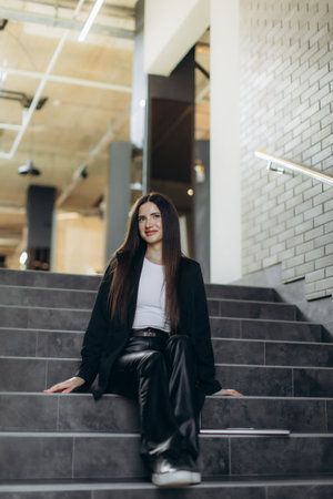Confident woman with long hair dressed in a chic outfit sitting on a staircase in a modern indoor space, exuding elegance and sophistication, showcasing contemporary fashion in an urban professional setting.の写真素材