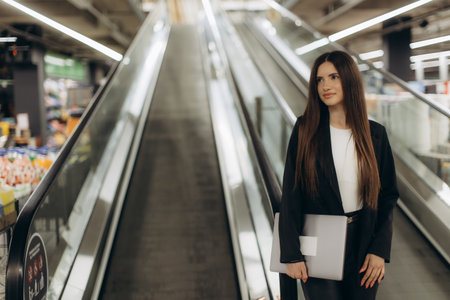 Professional young woman standing on an escalator, confidently holding a folder, symbolizing ambition and modern lifestyle in a commercial environment.の写真素材
