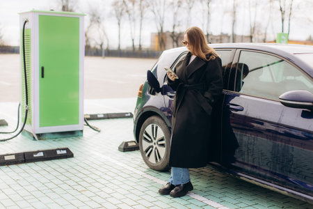 Woman standing next to a charging electric vehicle in a parking lot, connecting to technology for sustainable transportの写真素材