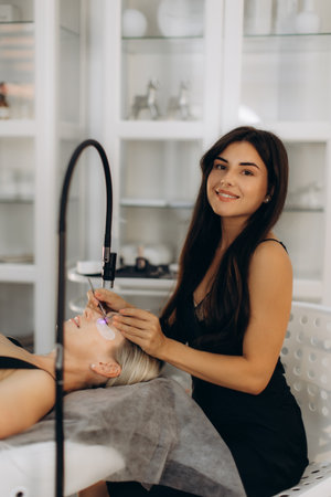 Professional beautician applying eyelash extensions to a woman's eyes using a special led lamp in a modern beauty salonの写真素材