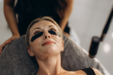 Woman lying down with under-eye patches, receiving led eyelash extensions from a beautician in a beauty salonの写真素材