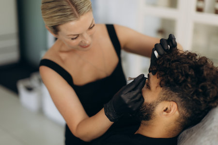 Beautician in black gloves performing a permanent makeup procedure on a man's eyebrows, providing cosmetic treatmentの写真素材