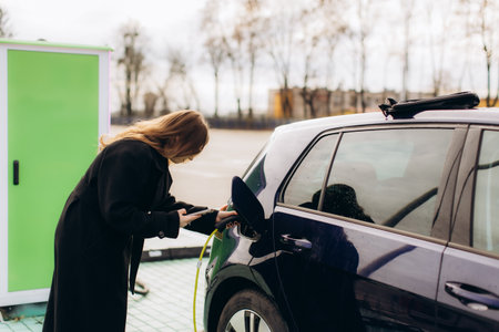 Woman standing by a blue electric vehicle, plugging in a charger and holding a smartphone, representing sustainable transportationの写真素材
