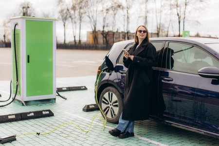 Woman standing next to her electric vehicle at a public charging station, checking her smartphone during the charging processの写真素材