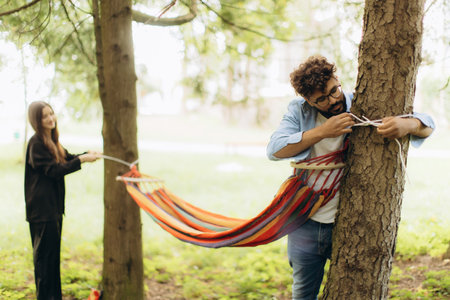 Young couple collaborating, tying colorful striped hammock to trees in a park, enjoying leisure outdoorsの写真素材
