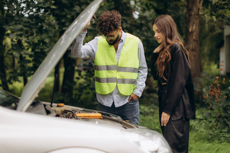 Young couple dealing with a broken-down car, the man checking the engine while wearing a safety vestの写真素材