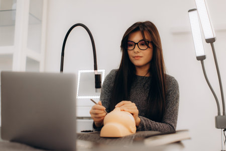 Young woman practicing permanent makeup on a mannequin head, watching an online lesson on her laptop...