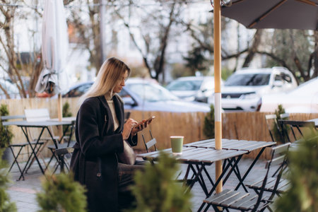 Young blonde woman using a mobile phone and drinking coffee at an outdoor street cafe, enjoying technology and urban lifestyleの写真素材