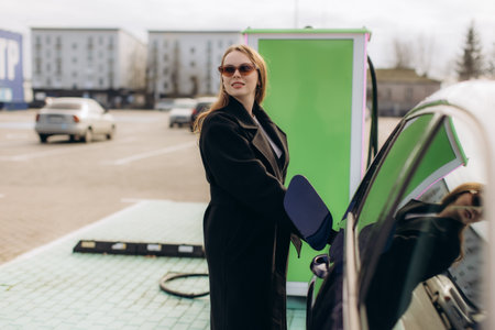 Woman standing next to electric car, plugging in charging cable at a public ev charging station, promoting sustainable transportationの写真素材