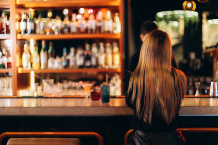 A woman with long blonde hair sits at a bar counter. A warmly lit collection of bottled beverages and prepared cocktails provides an ambient background, reflecting a cozy and social atmosphere.の写真素材