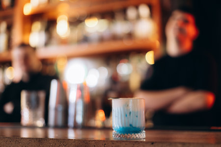 A close-up of a vibrant cocktail drink on a bar counter, surrounded by a warm bokeh light background and blurred bar staff in the distance, creating a lively and inviting ambiance.の写真素材