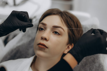 Professional beautician shaping young woman's eyebrows with threading tool in a beauty salon for grooming procedureの写真素材