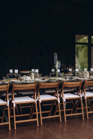 Outdoor long dining table prepared for a festive occasion or wedding, featuring wooden chairs,...