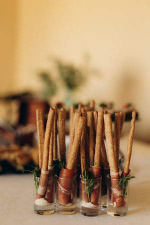 Breadsticks wrapped in thin slices of prosciutto and fresh greens, served in small glasses on a buffet tableの写真素材