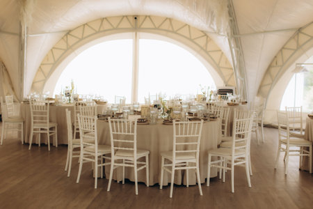 Elegant white tent prepared for a wedding reception or banquet, featuring round tables with formal...