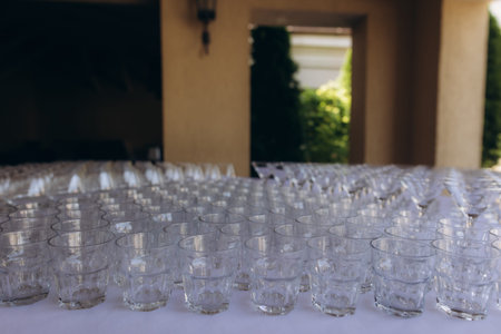 Rows of clean, empty glasses arranged on a white table, ready for drinks at an outdoor reception or...