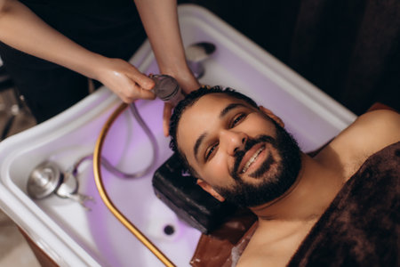 Smiling man enjoying a professional hair wash and relaxing head massage at a modern barbershop spa, feeling refreshed and pamperedの写真素材
