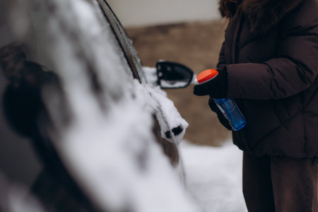 Person removing ice from a car window and side mirror during winter with a blue de-icer spray bottle