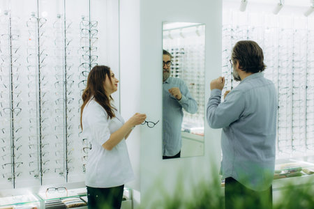 Optician assisting a male customer trying on eyeglasses frames in front of a mirror at an optical storeの写真素材