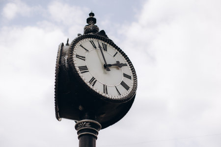 Ornate street clock with roman numerals, black hands and white face against a bright cloudy sky,...