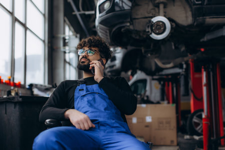 Male auto repair technician in overalls making a call, discussing car maintenance solutions inside a professional garageの写真素材