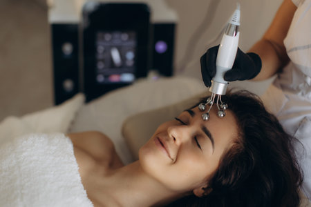 Woman relaxing on spa bed receiving microcurrent facial treatment for anti-aging skin rejuvenation,...