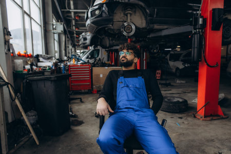 Male mechanic with a beard and glasses sitting on a chair looking up in a car service station, surrounded by tools and vehiclesの写真素材